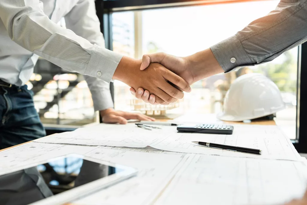 A general contractor shaking hands with a client over a desk covered in blueprints and commercial construction documents