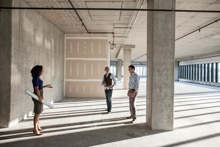 The interior of an unfinished empty building during a commercial renovation or remodel, where three construction professionals stand together holding blueprints